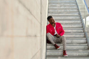 Junger Mann in roter Jacke und karierter Hose sitzt auf einer Treppe im Freien - AODF00243