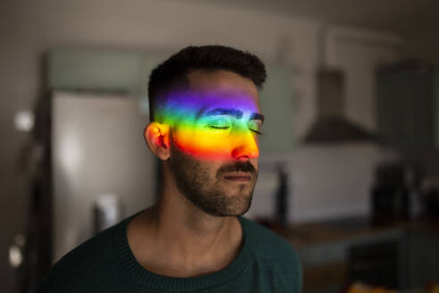 Regenbogenlicht trifft das Gesicht eines jungen Mannes, der mit geschlossenen Augen in einem Haus steht - MIMFF00390