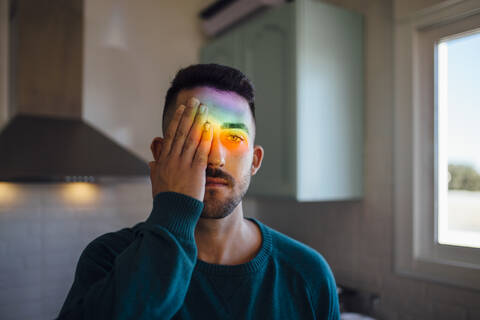 Rainbow light hitting face of young man covering one eye with hand stock photo