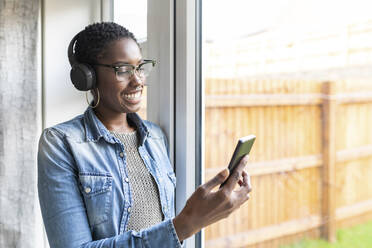 Lächelnde Frau mit Kopfhörern steht am Fenster und schaut auf ihr Smartphone - WPEF03876