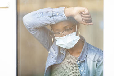 Sad woman in face mask leaning on window - WPEF03895