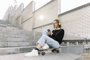 Junge Frau sitzt auf einem Skateboard und hört Musik im Freien - XLGF00981