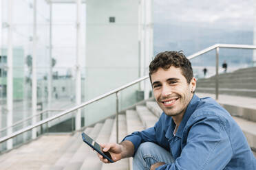 Porträt eines lächelnden jungen Mannes, der ein Smartphone im Freien hält - XLGF00987