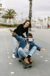 Frau schiebt Mann auf Skateboard sitzend mit Virtual-Reality-Brille - XLGF01020