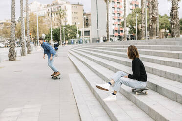 Frau sitzt auf einer Treppe und sieht einem Mann beim Skateboarden zu - XLGF01025