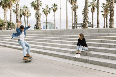 Junge Frau, die auf einer Treppe sitzt und einem Mann beim Skateboarden zusieht - XLGF01028