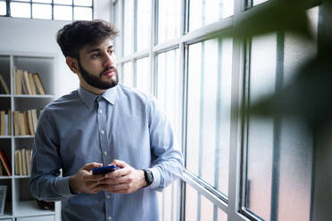 Junger Geschäftsmann, der sein Smartphone hält und durch ein Fenster in einem kreativen Büro schaut - GIOF10548