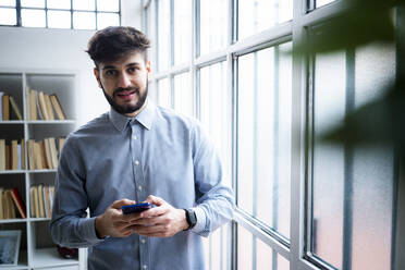 Portrait of young businessman holding phone, standing by window in creative office - GIOF10550