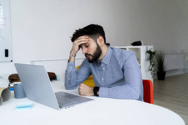 Tired businessman looking at laptop in creative office - GIOF10566