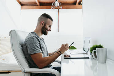 Lächelnder junger Mann, der zu Hause an Laptop und Mobiltelefon arbeitet - EBBF02120