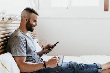 Lächelnder junger Mann, der zu Hause auf dem Bett sitzend an seinem Mobiltelefon arbeitet - EBBF02136