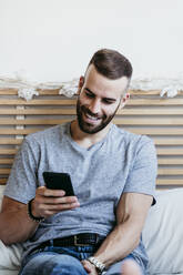 Lächelnder junger Mann, der zu Hause auf dem Bett sitzend an seinem Mobiltelefon arbeitet - EBBF02137