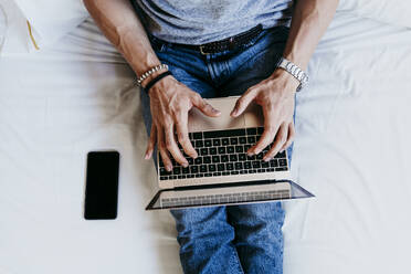 Blick von oben auf einen jungen Mann, der an einem Laptop und einem Mobiltelefon arbeitet, während er zu Hause auf dem Bett sitzt - EBBF02138