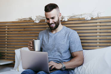 Lächelnder junger Mann arbeitet am Laptop, während er zu Hause auf dem Bett sitzt - EBBF02139