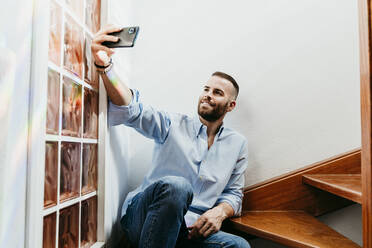 Smiling young man taking selfie while sitting on wooden stairs at home - EBBF02146