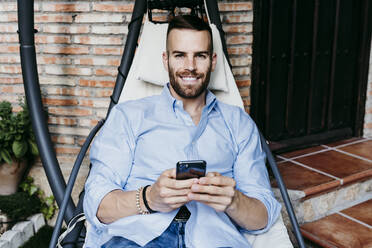 Smiling young man working with mobile phone while sitting in hammock on terrace - EBBF02155