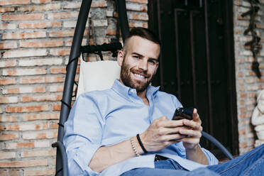 Lächelnder junger Mann, der mit seinem Handy arbeitet, während er in der Hängematte auf der Terrasse sitzt - EBBF02157