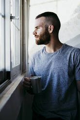 Young man holding cup of coffee while standing by window at home - EBBF02167