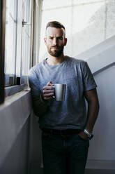 Young man holding cup of coffee while standing by window at home - EBBF02168