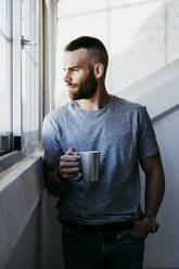 Junger Mann hält eine Tasse Kaffee in der Hand, während er zu Hause am Fenster steht - EBBF02169