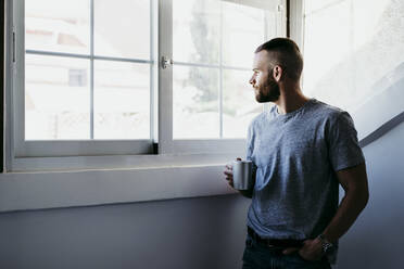 Junger Mann hält eine Tasse Kaffee in der Hand, während er zu Hause am Fenster steht - EBBF02170