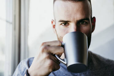 Young man holding cup of coffee while standing by window at home - EBBF02171