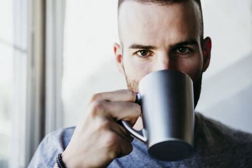 Junger Mann hält eine Tasse Kaffee in der Hand, während er zu Hause am Fenster steht - EBBF02171