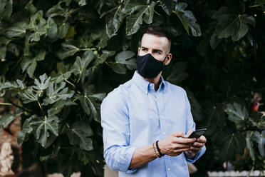 Junger Mann trägt eine Schutzmaske und benutzt ein Mobiltelefon - EBBF02175