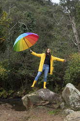 Carefree young woman wearing yellow raincoat holding umbrella while standing on rock in forest - KBF00651