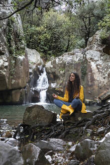 Junge Frau meditiert im Sitzen auf einem Felsen vor einem Wasserfall im Wald während der Regenzeit - KBF00662