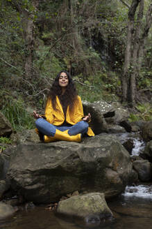 Female hiker wearing yellow raincoat meditating while sitting on rock in forest - KBF00663