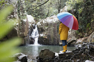 Frau mit Regenschirm schaut auf einen Wasserfall und steht auf einem Felsen im Wald - KBF00664