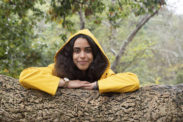 Lächelnde Wanderin in gelbem Regenmantel, die sich im Wald an einen Baumstamm lehnt - KBF00665