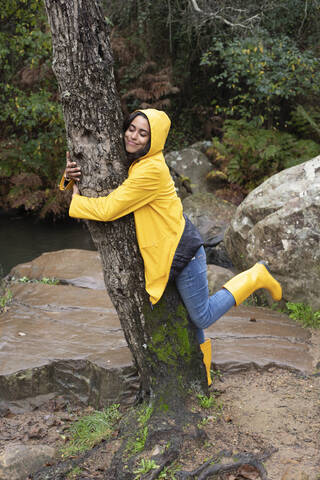 Happy female hiker wearing yellow raincoat embracing tree in forest stock photo