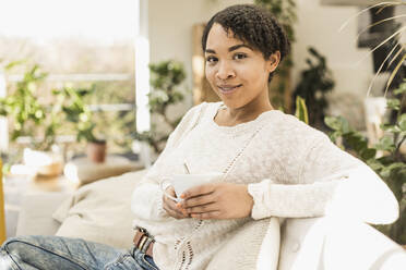 Lächelnde junge Frau hält eine Kaffeetasse, während sie zu Hause auf dem Sofa sitzt - UUF22582