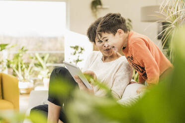 Mother showing digital tablet to son while sitting at home - UUF22596