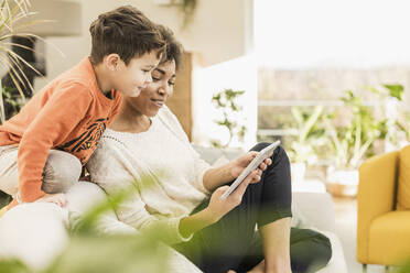 Frau zeigt einem Jungen zu Hause sitzend ein digitales Tablet - UUF22597