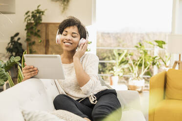 Junge Frau mit Kopfhörern, die Musik hört, während sie ein digitales Tablet benutzt, das zu Hause steht - UUF22603