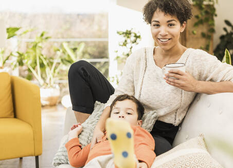 Frau mit Kaffeetasse, die lächelnd neben einem Jungen sitzt, der sich zu Hause auf dem Sofa ausruht - UUF22605