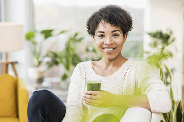 Young woman wearing eyeglasses smiling while sitting with juice at home - UUF22607