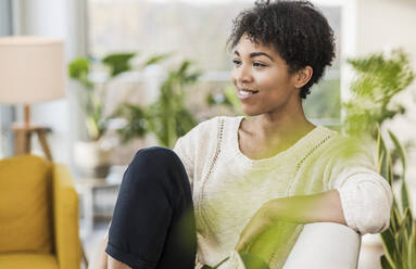 Smiling woman looking away while sitting on sofa at home - UUF22609