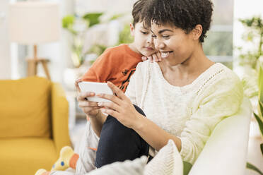 Mutter, die ein Mobiltelefon benutzt, während sie mit ihrem Bruder zu Hause auf dem Sofa sitzt - UUF22610