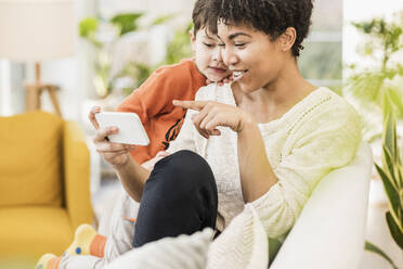 Lächelnde Mutter zeigt ihrem Sohn das Handy, während sie zu Hause sitzt - UUF22611