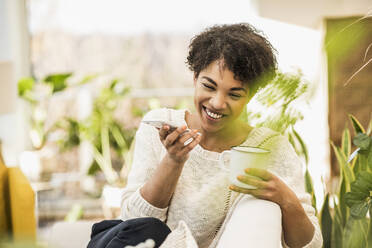 Smiling woman with coffee cup talking on mobile phone while sitting at home - UUF22617