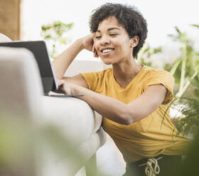 Young woman smiling while using digital tablet sitting at home - UUF22620