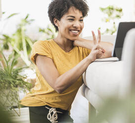 Smiling woman showing peace gesture on video call through digital tablet while sitting at home - UUF22624