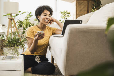 Junge Frau mit Mobiltelefon, die zu Hause sitzend auf einem digitalen Tablet spricht - UUF22626