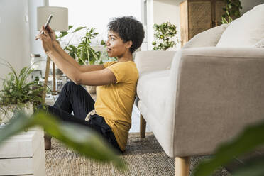 Junge Frau, die ein Selfie mit ihrem Mobiltelefon macht, während sie zu Hause auf dem Sofa sitzt - UUF22628