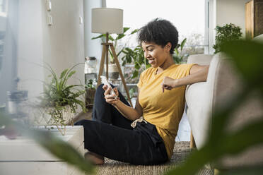 Woman smiling while using mobile phone sitting at home - UUF22629