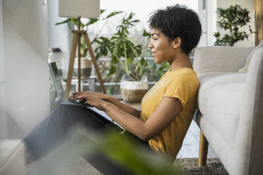 Young woman using digital tablet while leaning on sofa at home - UUF22632